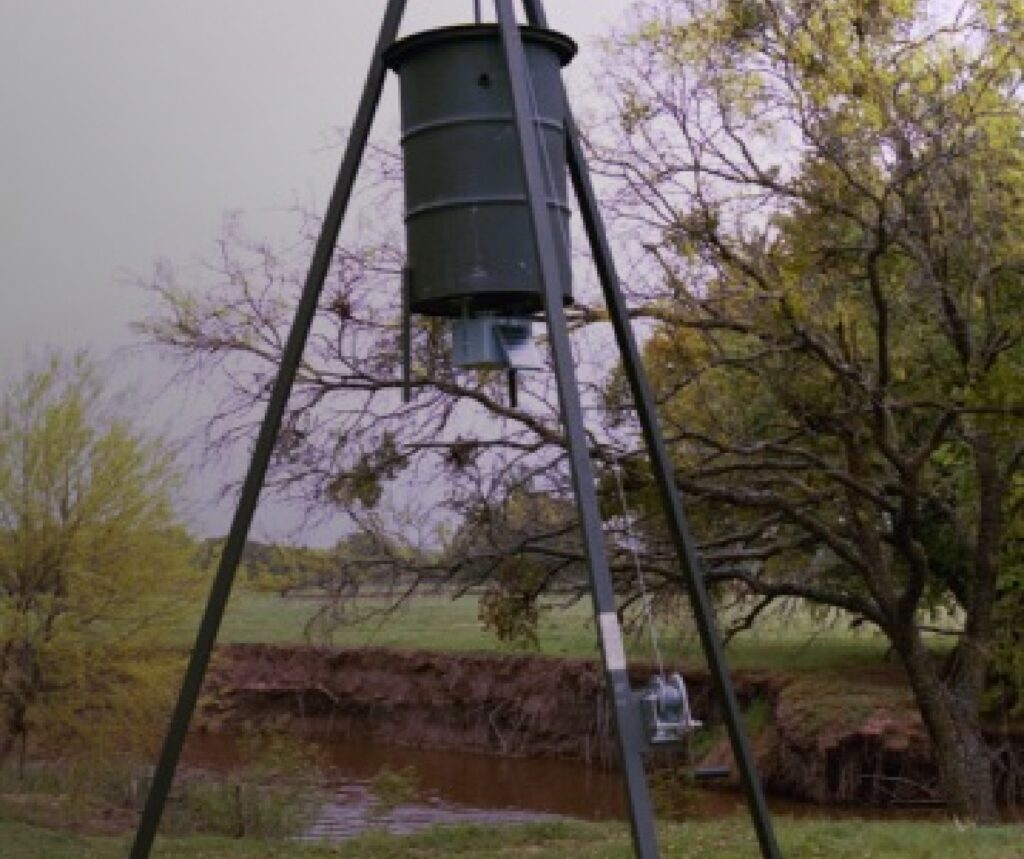 Bird feeder tower with a barrel attached to it on a tripod next to a stream